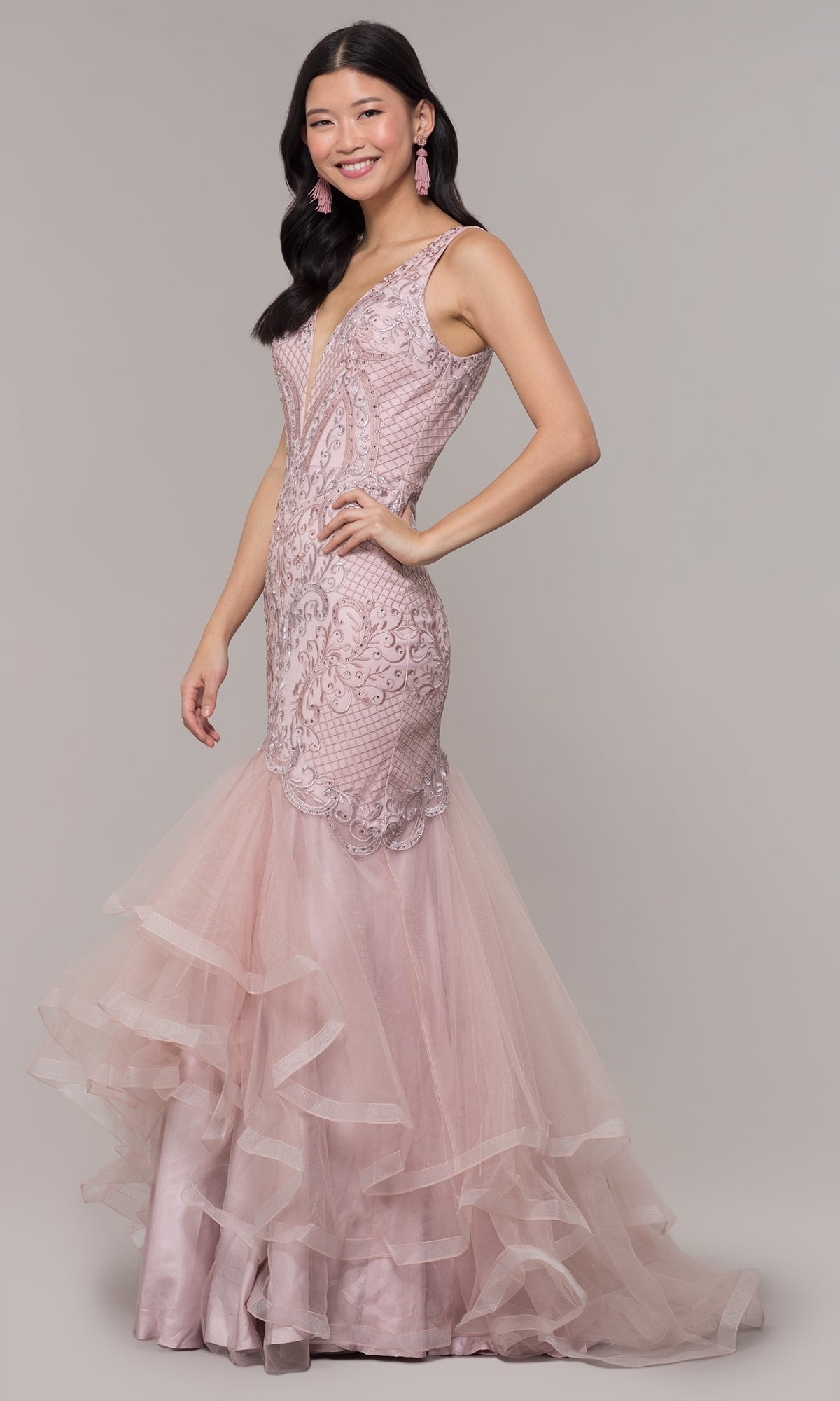 A woman wears the Embroidered V-Neck Mermaid Formal Prom Dress 8320, a sleeveless light pink gown with lace, tulle, and a deep V-neckline, as she smiles against a plain background.